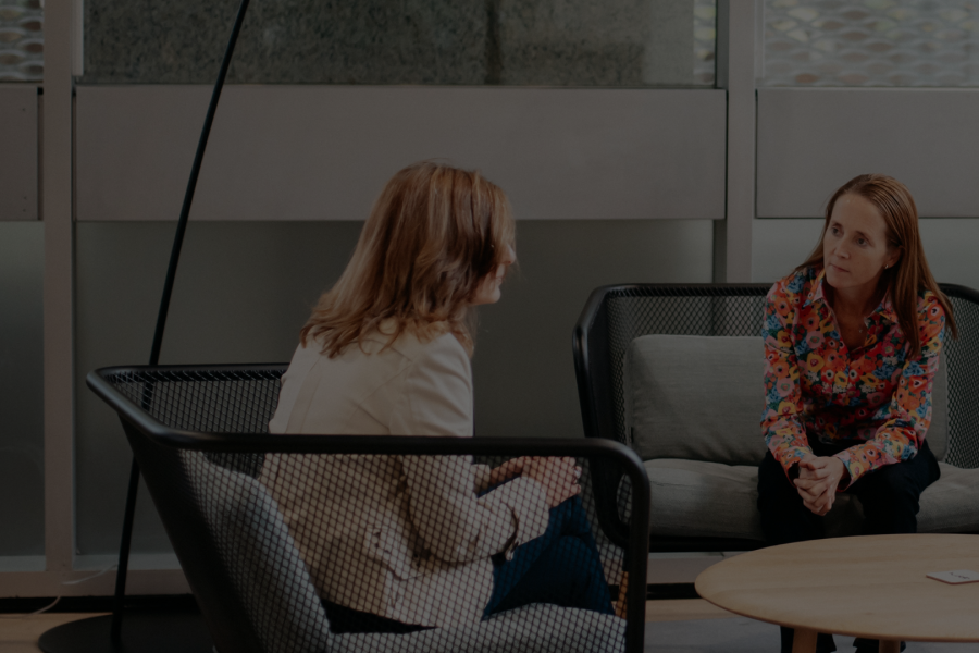 Two women sit on chairs leaning in as if in for a business support discussion