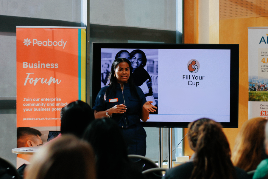 Woman stands in front of a tv screen presentation with rows of seated audience looking at her.